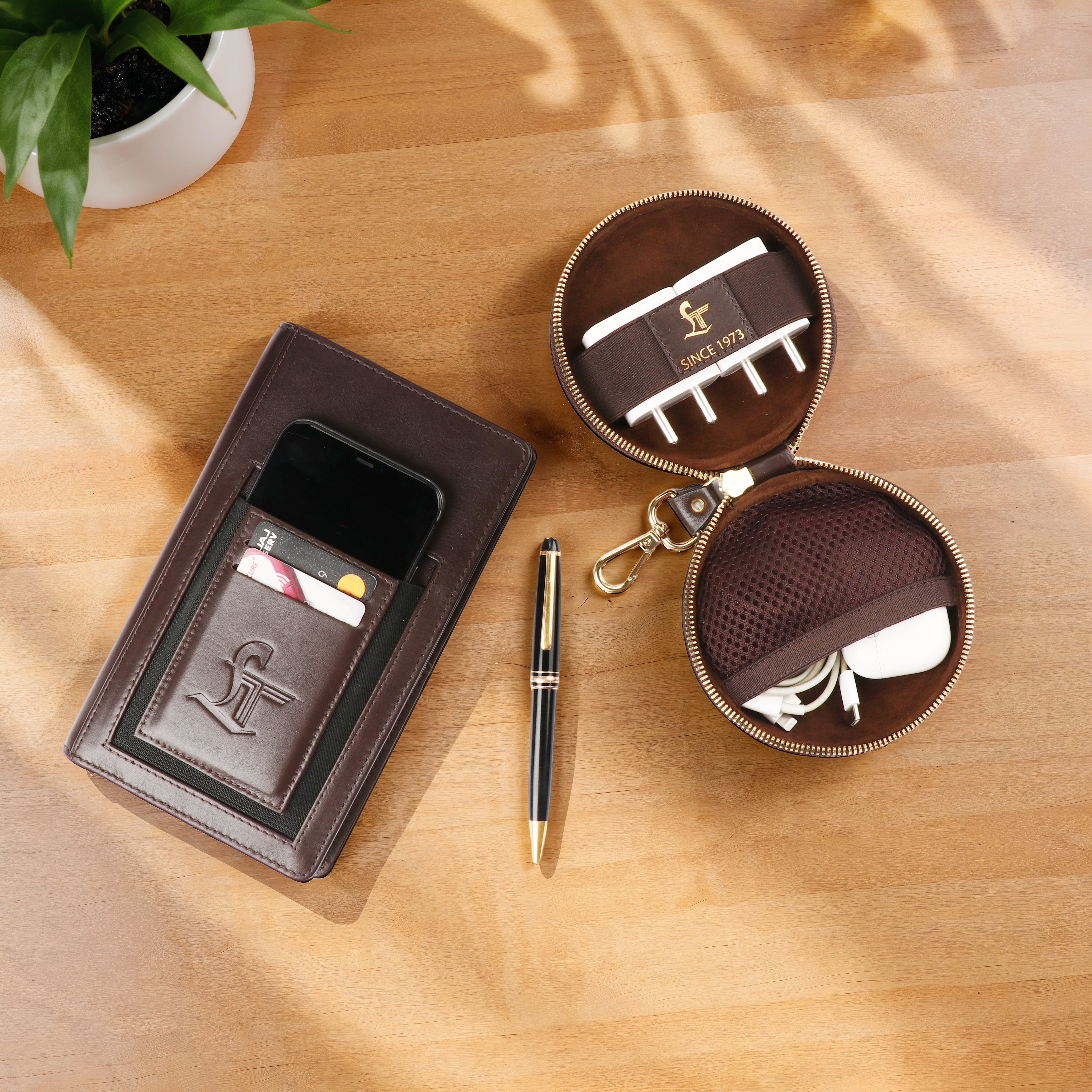 Brown leather wallet, round key case, and pen on a wooden surface with a plant in the corner.